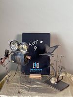 Photo showing both metal sculptures side by side on a cloth-covered surface with a backdrop sign reading 'LOT 9 THE NEXT STEP AWARD'