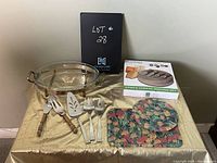 Clear oval Pyrex glass baking dish with metal stand, cheese set in box, floral glass trivet and rectangular mat, and serving cutlery with wood and white handles arranged on gold fabric