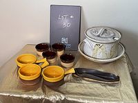 Photo showing a full view of the ceramic soup tureen with lid and ladle alongside four yellow onion soup bowls and five brown ramekins arranged on a gold-colored surface.