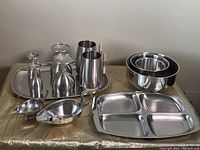 Photo showing the complete set of stainless steel dishes including trays, goblets, mugs, bowls, and other serveware pieces, arranged on a gold fabric surface.