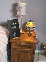 Two table lamps on wooden nightstand. Shorter lamp has faux Tiffany stained-glass shade, bronze base. Taller lamp has slender metal base and white fabric shade with bow.
