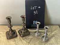 Two pairs of metal taper candle holders displayed on a table: two larger with square bases and two smaller with round bases, showing finish and condition.