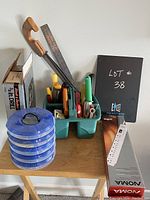 Photo of assorted hand tools organized in a green plastic caddy, a wooden handled hand saw, a metal ruler, a Blue five-drawer plastic organizer stack, boxed Black and Decker drill and boxed NOMA power strip all on a wooden table.