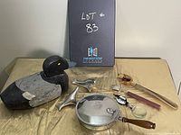 Lot 83 items including carved wooden duck, metal pan, dolphin sculptures, bed warmer, magnifying glass, pipe tool, and decorative smoking pipe on a yellow tablecloth with black board in background.