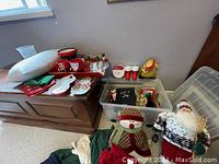 Overview of lot showing various aspects of Christmas decor and serving items on chest and floor including figurines, mugs, and boxes