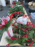 Top view of table displaying artificial lighted garland with pinecones and red berries, gift box with ribbon and candy cane picks, wreath with red berries and pinecones, and additional red ribbons