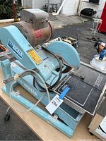 Side view of the tile saw showing blue painted body, motor on top labeled Ricker, circular saw blade, and table surface for cutting.