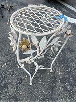 Side angle showing white wrought iron plant stand with flower and leaf decorations and lattice top surface, placed on asphalt ground.