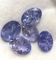 Close-up of five oval faceted violet-blue tanzanite gemstones on white cushioned background, showing clarity and color variations.