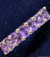 Close-up angled view of silver ring band set with seven faceted amethyst gemstones in prong settings against a dark fabric background.