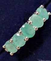 Close-up view of silver ring with six oval faceted emeralds showing prong setting and gemstone details.