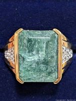 Front view of the rectangular green emerald set in gold-tone prongs with white topaz accents on each side, mounted on a silver ring band, against dark blue background.