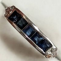 Top angled close-up of silver ring showing five square-cut blue stones and small round clear accents on both ends.