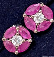 Close-up front view of the pair of silver earrings with pink ruby and clear stones on a dark fabric background.