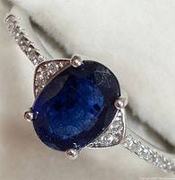 Close-up photo of silver ring with oval blue sapphire and cubic zirconia accents against white background.