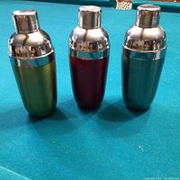 Front angled view of three cocktail shakers in green, red, and blue with shiny silver caps, standing upright on blue felt surface.