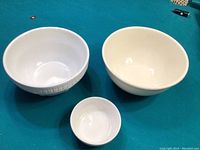 Three white ceramic bowls of varying sizes placed on green surface: two larger bowls with one showing embossed pattern and one smaller plain bowl.