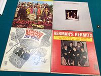 Four vinyl LP records laid out on a flat surface. Top left is The Beatles 'Sgt. Pepper's Lonely Hearts Club Band' with colorful iconic cover art. Top right is The Beatles 'Parlophone Mono' album with grey cover and small inset photo. Bottom left is Herman's Hermits 'On Tour' album with white illustrated whimsical cover. Bottom right is Herman's Hermits self-titled album with red cover and group photo.