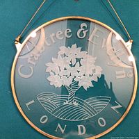 Front view of round etched glass sign with Crabtree & Evelyn logo and London text, showing etched tree design and metal rim.