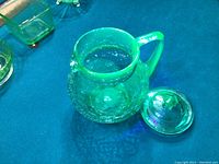 Crackle uranium glass jug with lid displayed on a blue surface, showing the jug open with the lid placed beside it, exhibiting the textured crackle glass pattern in natural light.