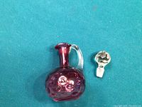 Front view of the cranberry glass cruet with its stopper placed beside it on a blue background. The cruet is bulbous with embossed circular patterning and a clear handle.