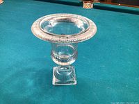 Clear glass urn vase photographed upright on a teal felt surface, showing overall shape and design details including flared rim, rounded midsection, and square base.