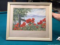 Framed watercolour painting depicts a landscape scene with vibrant red poppies in front, a fence post, trees, water, and mountains.