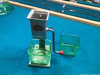 Metal ice crusher marked 'Lightning Ice Cube Breaker' with attached uranium glass bowls on blue billiard table.