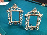 Two matching vintage metal picture frames on a table, showing full front views and stand supports.