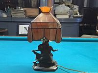 Side photo of stained glass lamp showing orange stained glass shade with black leading and metal figurine base of a woman in a flowing gown.