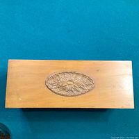 Closed wooden trinket box showing the simple rectangular shape with an ornate oval floral carved design on the lid.