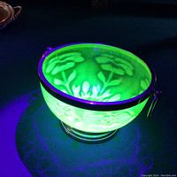 Uranium glass footed bowl glowing bright green under UV light showing floral design and metal rim with handles