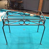 Front angle view of the wrought iron plant stand showing the decorative scroll design, rectangular shape, and slight leg curve.