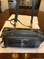 View of the front of the black leather handbag showing the front suede pocket with a strap detail and gold-tone hardware attaching the shoulder strap.