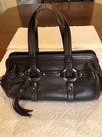 Front and side view of brown pebbled leather purse showing dual handles, decorative straps with gold-tone rings and rivets, leather tassel zipper pull.