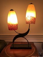 Front view of the lit lamp showing dual glass shades illuminated with warm yellow light and multi-color speckled gradient on bottom portion of shades.