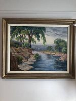 Frontal view of the framed oil painting depicting a river landscape with trees and rocky banks.