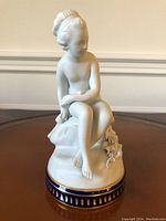 Front side view of white bisque porcelain figurine of young girl sitting on rock with flower detail and blue gold base
