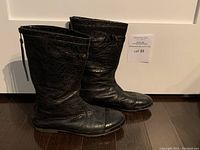 Side view of a pair of black leather mid-calf boots with textured leather, zipper on the back, and decorative strap with metal studs near top.