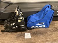 Pair of Bauer black and white ice hockey skates with TUUK blades and blue Bauer carrying bag on floor.