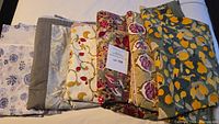 Group of seven folded Belle Provence cotton tablecloths in various floral and geometric prints arranged on a flat surface.