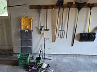 Wide view of gardening tools hanging on wall including shovels, rakes, pitchfork, and a snow shovel; also crates and step ladder on floor.
