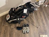 Overview of Wilson Staff golf bag with clubs inserted, shoes and sunglasses case arranged in front.