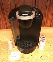 Front view of black Keurig B40 coffee maker with water reservoir, drip tray, two Donut Shop K-Cup pods and one reusable pod filter on table.