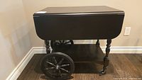 Wooden tea trolley with drop leaves folded down showing large spoked wheels on one side and smaller caster wheels on the other