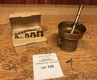 Full view of the pharmacy apothecary weight set in plastic case with brass weights, next to the bronze mortar and pestle on a wooden surface.