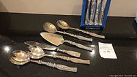 Full view of six silver serving utensils with detailed beaded handles, laid out on black surface.