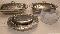 Overview showing three silver plated covered dishes with ornate grapevine borders and handles alongside six crescent shaped clear crystal plates.
