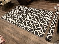 Pair of black and white outdoor rugs laid flat showing geometric interlocking ovals pattern on wood floor.