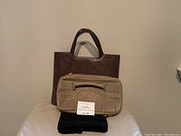 Two purses, the brown Levenger tote in back and the tan textured Alexander Wang clutch in front on top of black dust bag and with auction lot card
